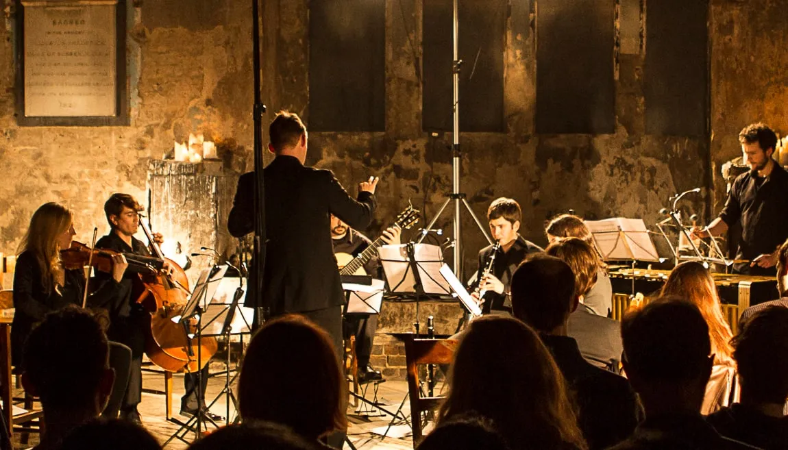 Photographed, from the back of the audience, a man conducts a small ensemble in low lighting