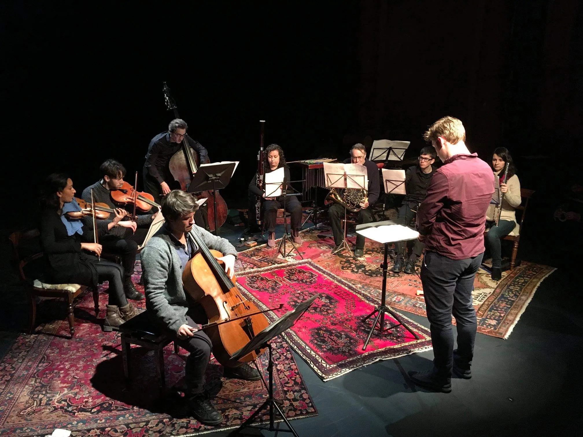 A close-up shot of a male solo cellist, ensemble of seven musicians playing stringed and woodwind instruments in a semicircle behind him, and conductor Jon Hargreaves standing in the middle in partial profile to the camera. They are in rehearsal on stage, dressed informally, with a black stage with layered Persian rugs.