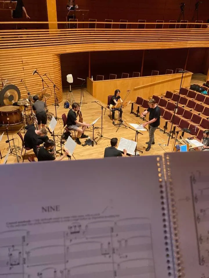 Photo of conductor Jon Hargreaves and spatially distanced musicians from Octandre Ensemble recording a piece of music in a timber-clad modern concert hall. The photo is taken from the perspective of one of the musicians, playing from a balcony above the stage, and their music fills the bottom half of the frame.
