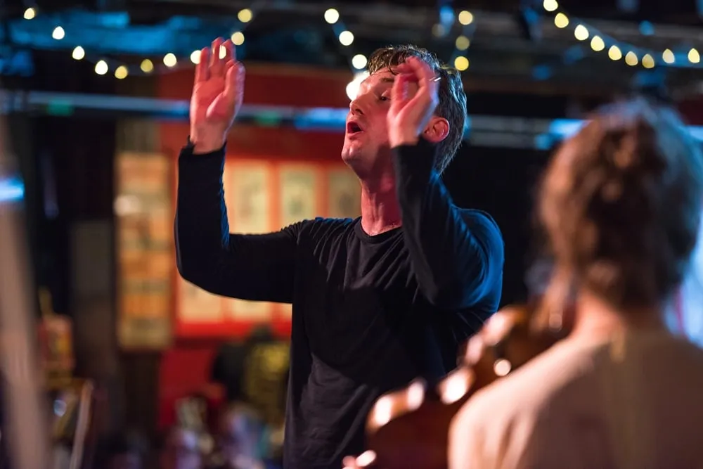 A closeup photo of Jon Hargreaves conducting the Nevis Ensemble in a dark space, lit by fairy lights.His arms are raised, and he wears a black shirt. in the foreground is the back of a violinist, with her hair pulled up into a chignon.