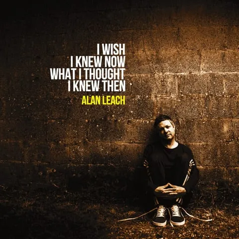 Singer Alan Leach, a white man with short hair, dressed in a black tracksuit with stripes, and wearing sneakers with the laces undone. He is crouched on his heels, hugging his knees leaning against an urban brick wall under lamplight.