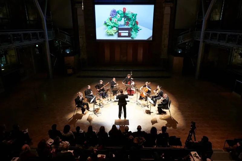 A symmetrical photo of 7 musicians from the London Symphony Orchestra in a semicircle around a brightly lit stage, with Jon hargreaves conducting at the centre (with his back to the camera). The photo is taken from the middle of the audience. The musicians are dressed in black clothing, and the mood is formal and professional.