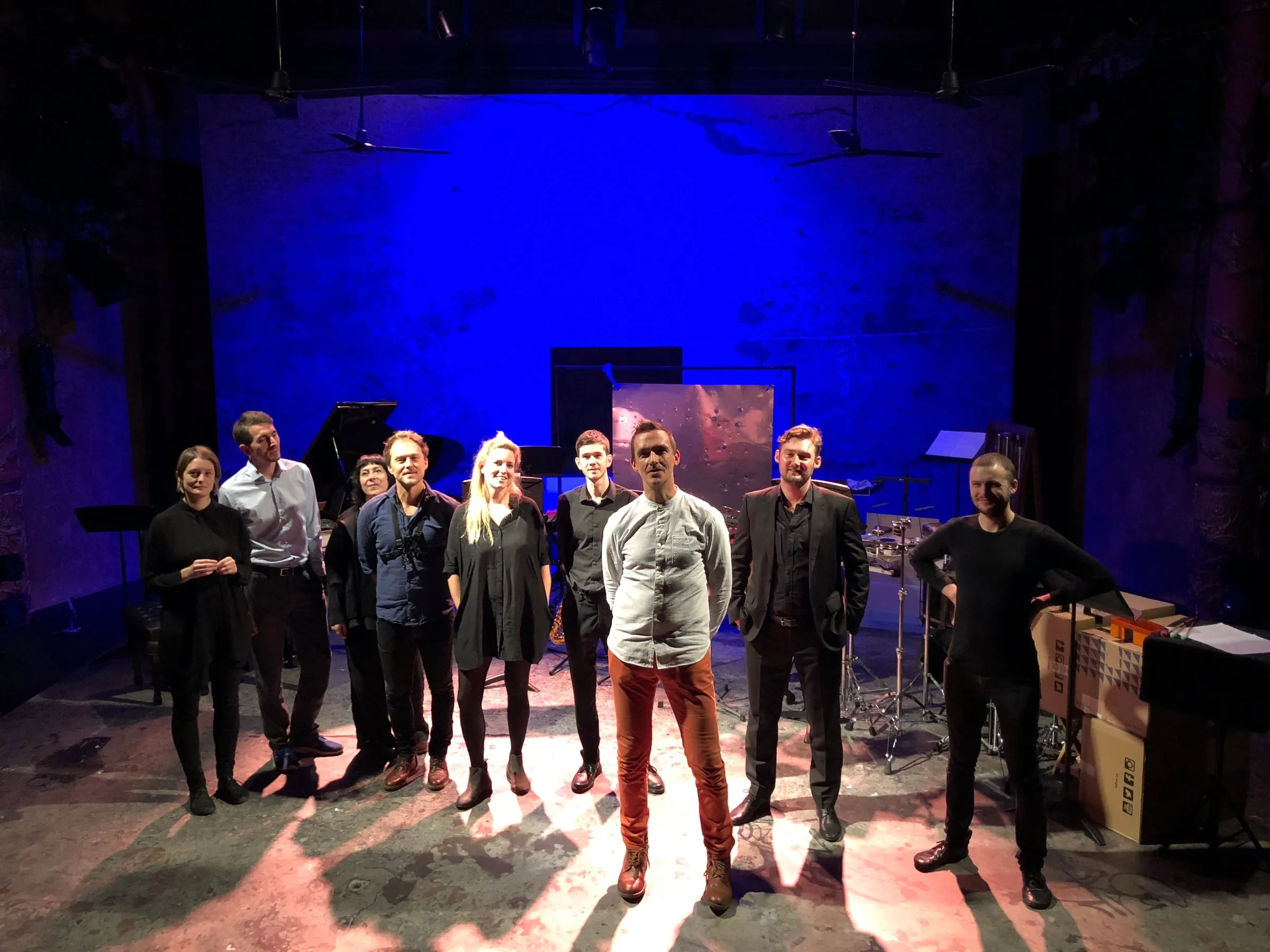 Eight musicians stand in a semicircle, infront of a battery of interesting percussion instruments. in front of them is composer Rolf Hind, in a white shirt and terracotta-coloured trousers. The backdrop is a bright blue, and the stage is brightly lit. They look directly at the camera.