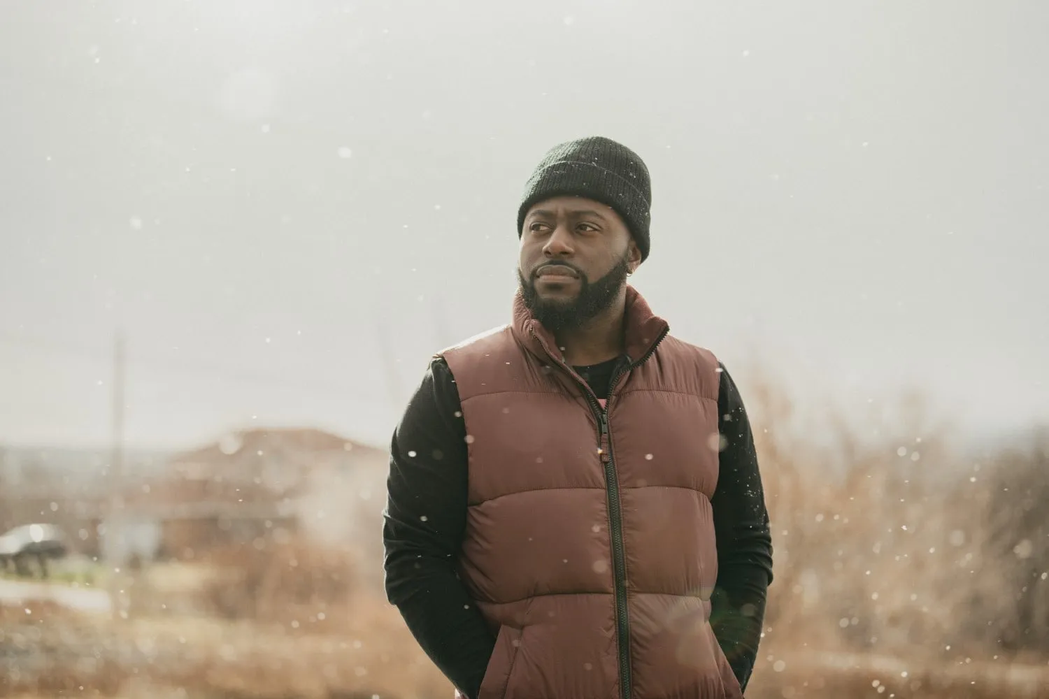 Closeup of African-Canadian hip-hop artist Keonte Beals. He stands in a rural setting, with snow swirling. He wears a brown puffy vest over a black jumper, with a black beanie, and has a clippered beard and moustache. He looks slightly towards the left frame of the photo.