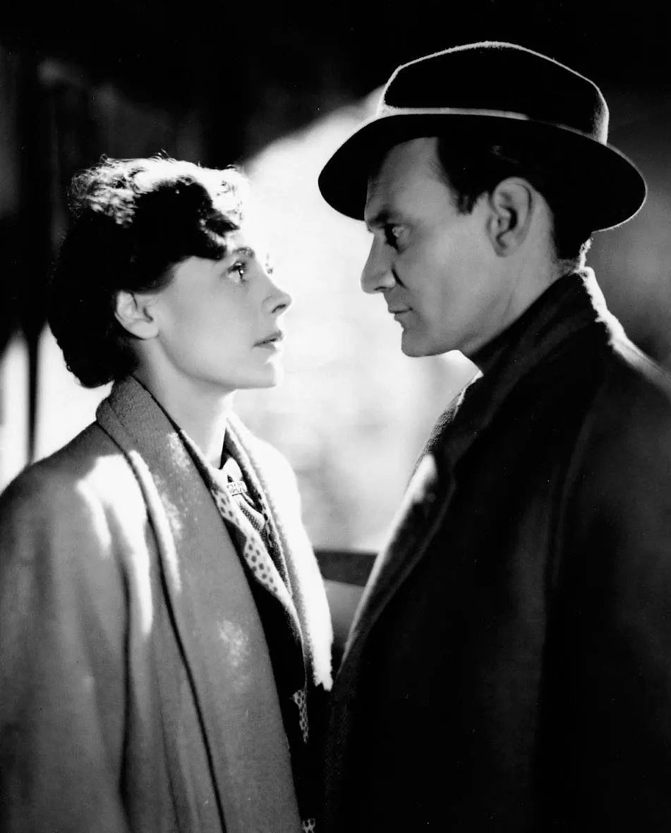 Camera still from the David Lean film "Brief Encounter". A black and white photo of a man and woman, both white and middle-aged, looking into each other's eyes, backlit by street lamps/