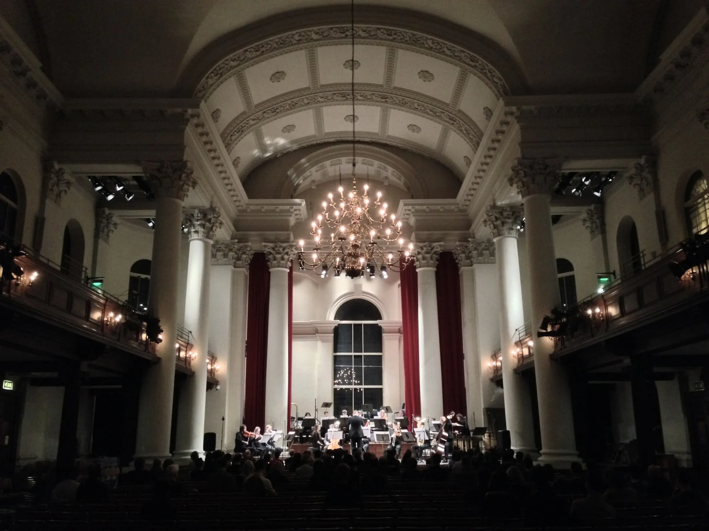 Image of the Octandre Ensemble in concert, in St John's Smith Square, London. the stage has a domed ceiling, draped red velvet panels either side of the performers, and a large chandelier above the stage.