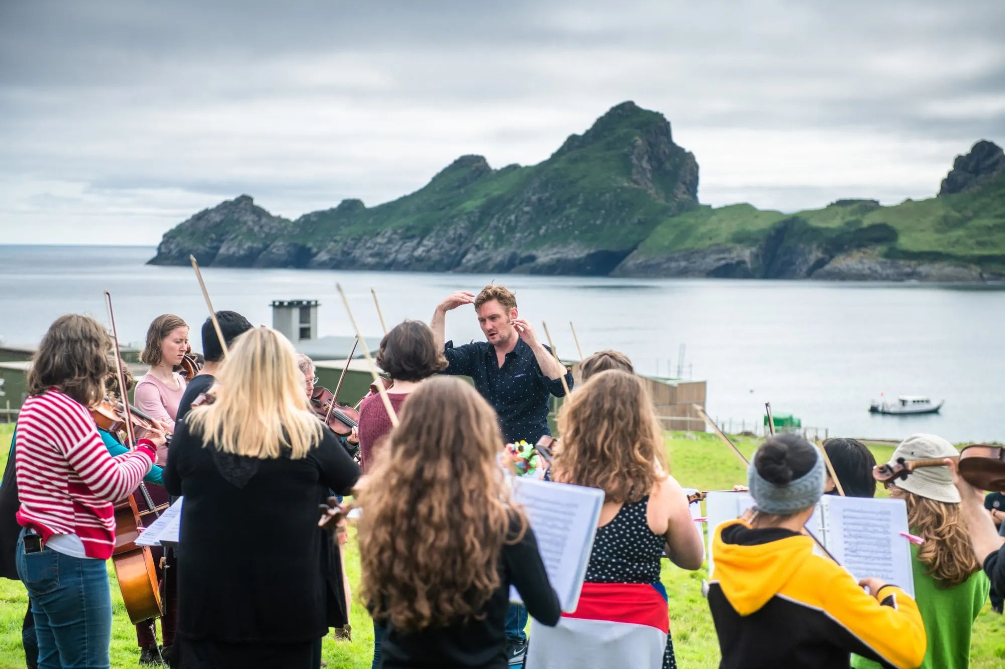A photo of the Nevis Ensemble musicians, with their back to the camera, playing with Jon Hargreaves conducting (facing the camera), on the Island of Hirta in the St Kilda archipelago. They are outdoors, with an inlet from the Atlantic ocean, and a rugged peninsula behind them. They are dressed in brightly coloured clothing.