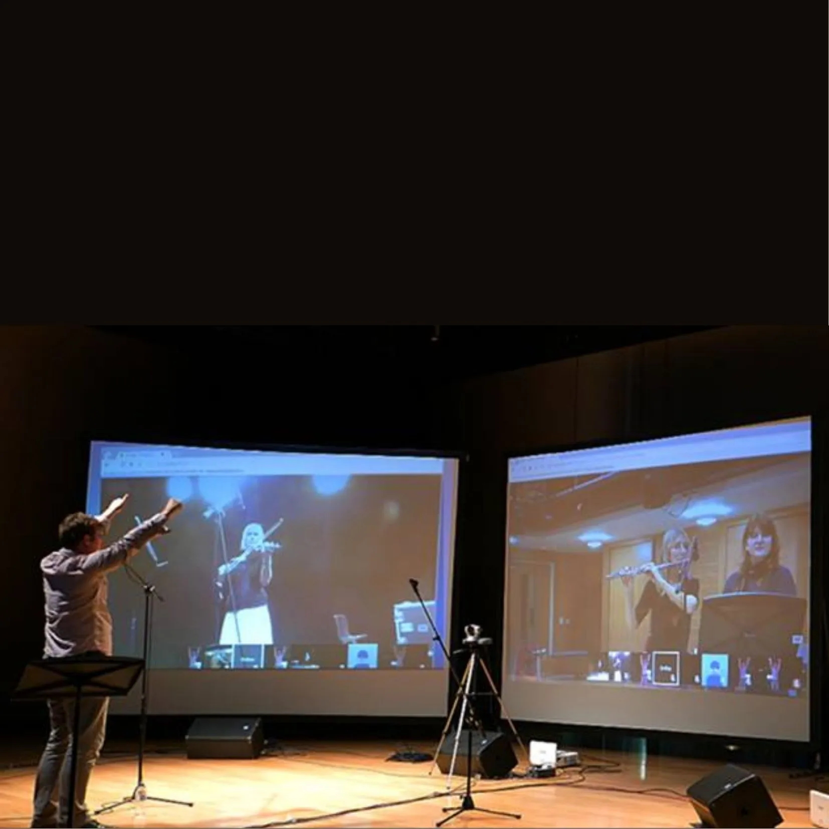 photo of Jon Hargreaves conducting into a camera, with live projections of different groups of musicians on three screens around him. Taken during the Online Orchestra research project at Falmouth University.