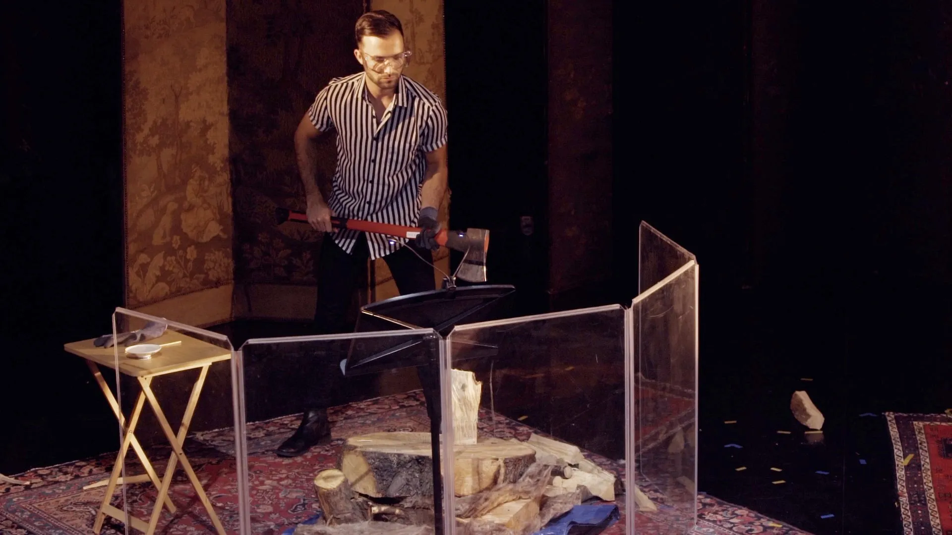 Caucasian man in striped shirt and black jeans, with smooth black hair, wearing safety goggles. He is chopping wood behind a plexiglass safety screen, on stage as part of a musical performance.