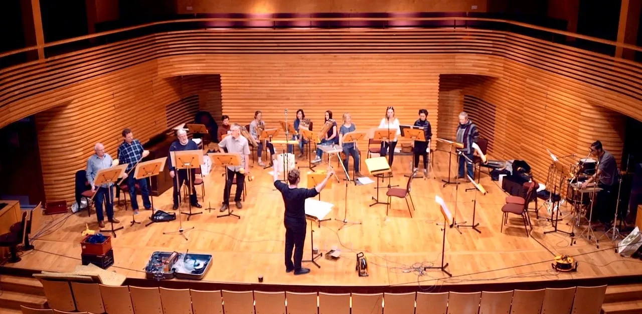 An image of the Octandre Ensemble recording on a modern, timber-lined concert platform. There are 3 groups of 4 players playing a large assortment of instruments in a large semi-circle around the stage, plus Jon Hargreaves conducting in the middle. The mood is informal, but busy.